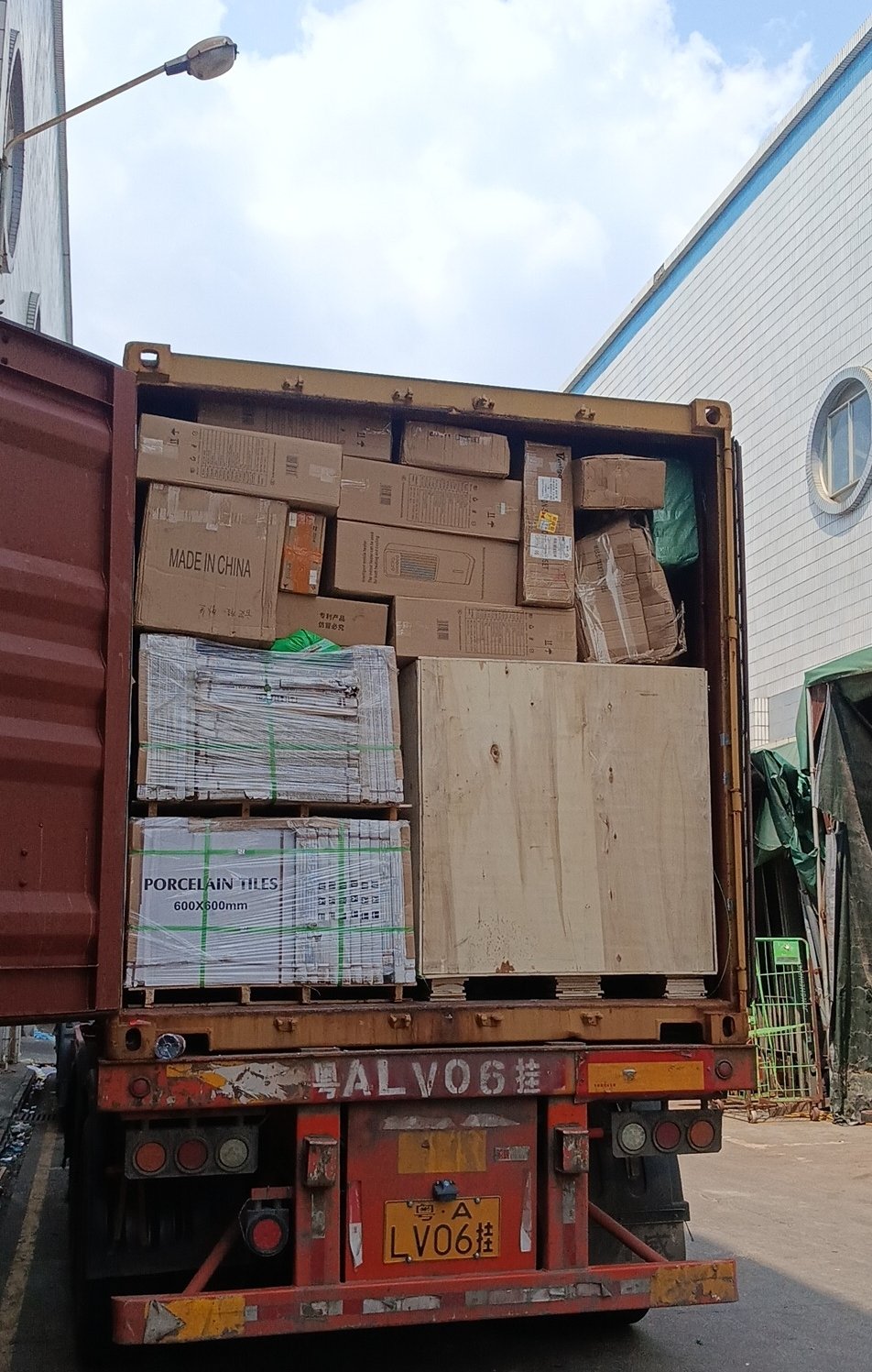 A loaded shipping container photographed from the open doors at a warehouse, showing cartons, stretch-wrapped pallets of porcelain tiles and a plywood-cased unit braced across the load, with the trailer unit number on the rear doors